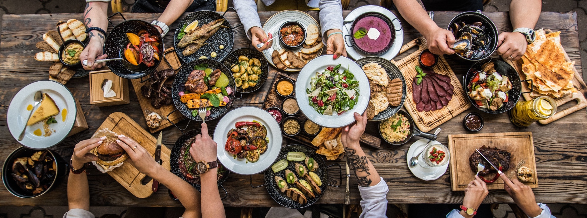 A group of people seated around a wooden table share a variety of farm-to-table dishes, including meats, salads, soups, grilled vegetables, bread, and drinks. TheCentralCoast.com