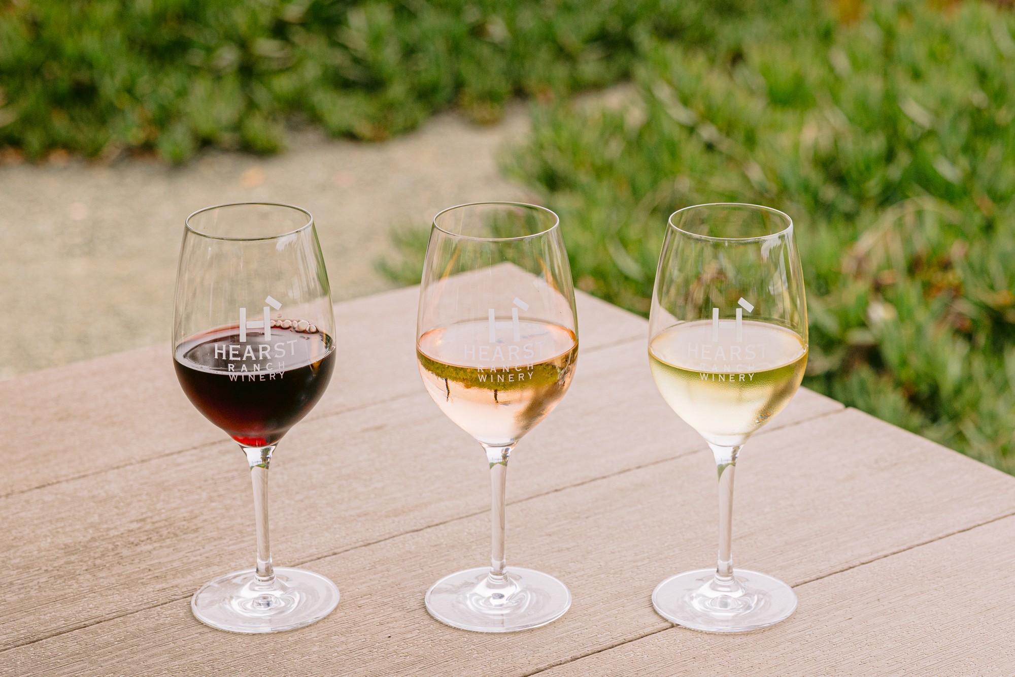 Three wine glasses—red, rosé, and white—sit on a wooden surface outdoors with green grass in the background, capturing the relaxed vibe of Central Coast Outdoors. TheCentralCoast.com