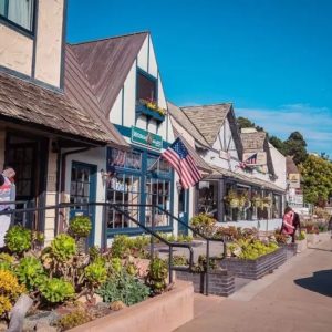 A row of quaint shops, including a charming seafood restaurant and oyster bar, with flower boxes and an American flag, line a sunny sidewalk where a few people stroll past parked cars. TheCentralCoast.com