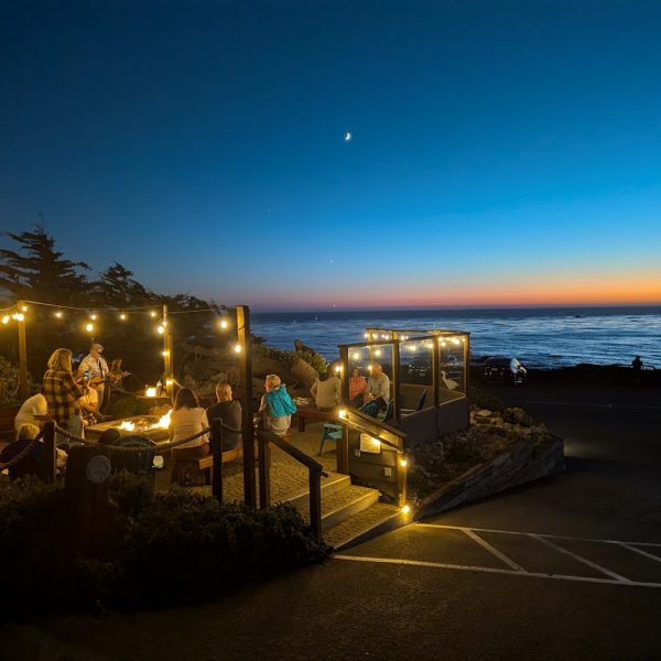 People dine outdoors at sunset by the ocean at Sea Chest Oyster Bar, with string lights illuminating the seating area and a crescent moon visible in the sky. TheCentralCoast.com