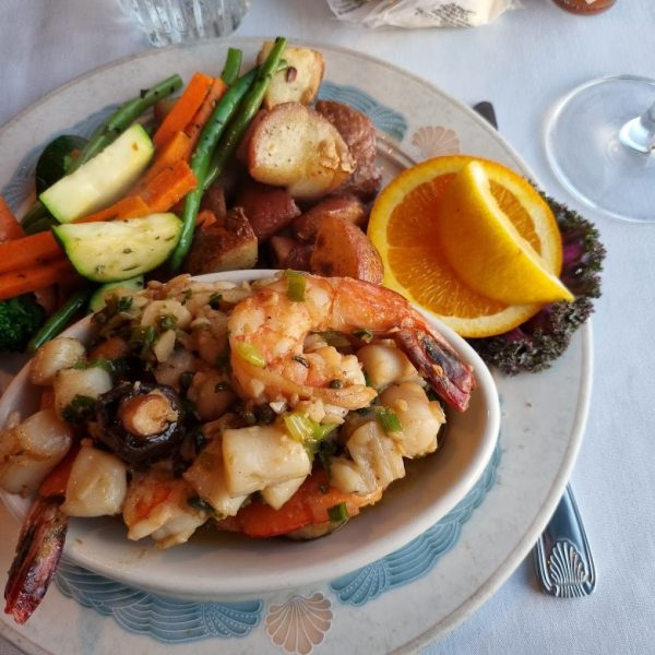 A plate with mixed vegetables, roasted potatoes, orange slices, and a bowl of shrimp and scallop stir-fry, set on a white tablecloth beside a glass of white wine at the Sea Chest Seafood Restaurant. TheCentralCoast.com