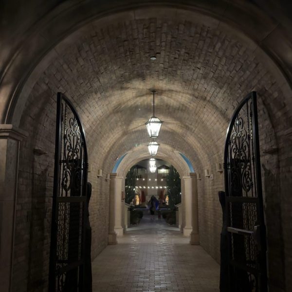 A brick archway corridor with ornate black gates, illuminated by hanging lanterns, leads to a courtyard at Central Coast Zoos where plants and people are visible in the distance. TheCentralCoast.com