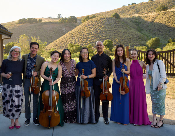 Nine people, some holding string instruments, stand in a row outdoors in front of hills at Central Coast Zoos, smiling at the camera on a sunny day TheCentralCoast com